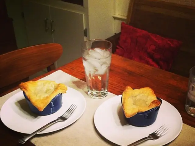 In the center of the image there is a dining table. There are plates, forks, cups, glasses and some...